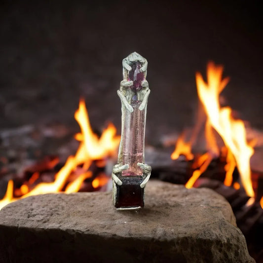 Handcrafted gemstone pendant with sculpted metal details on a rock, fire in the background