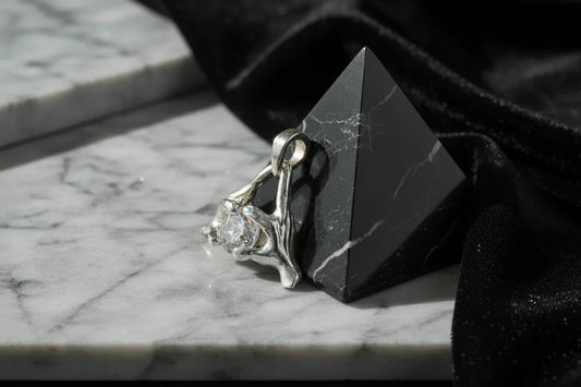Handcrafted silver pendant with clear gemstone on marble surface beside black pyramid sculpture