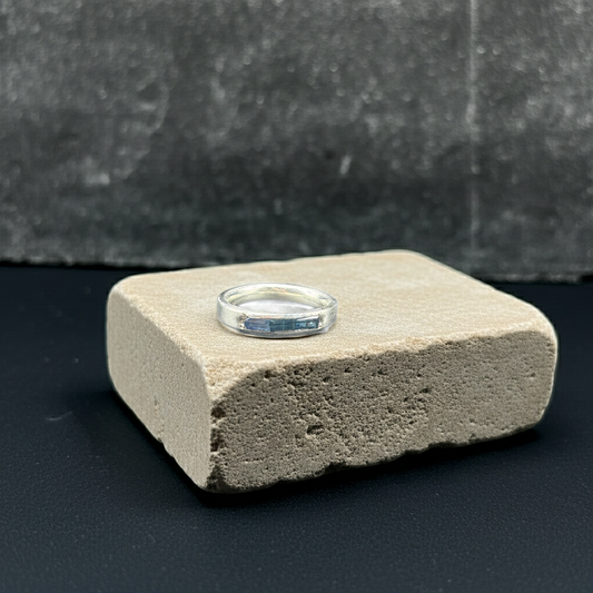 A thin silver band ring with a blue gemstone stack , displayed on a textured beige stone block against a dark gray background.