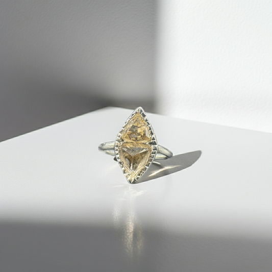 A silver ring featuring a marquise-cut yellow gemstone in a decorative bezel setting, displayed on a white surface with dramatic side lighting and a soft reflection.