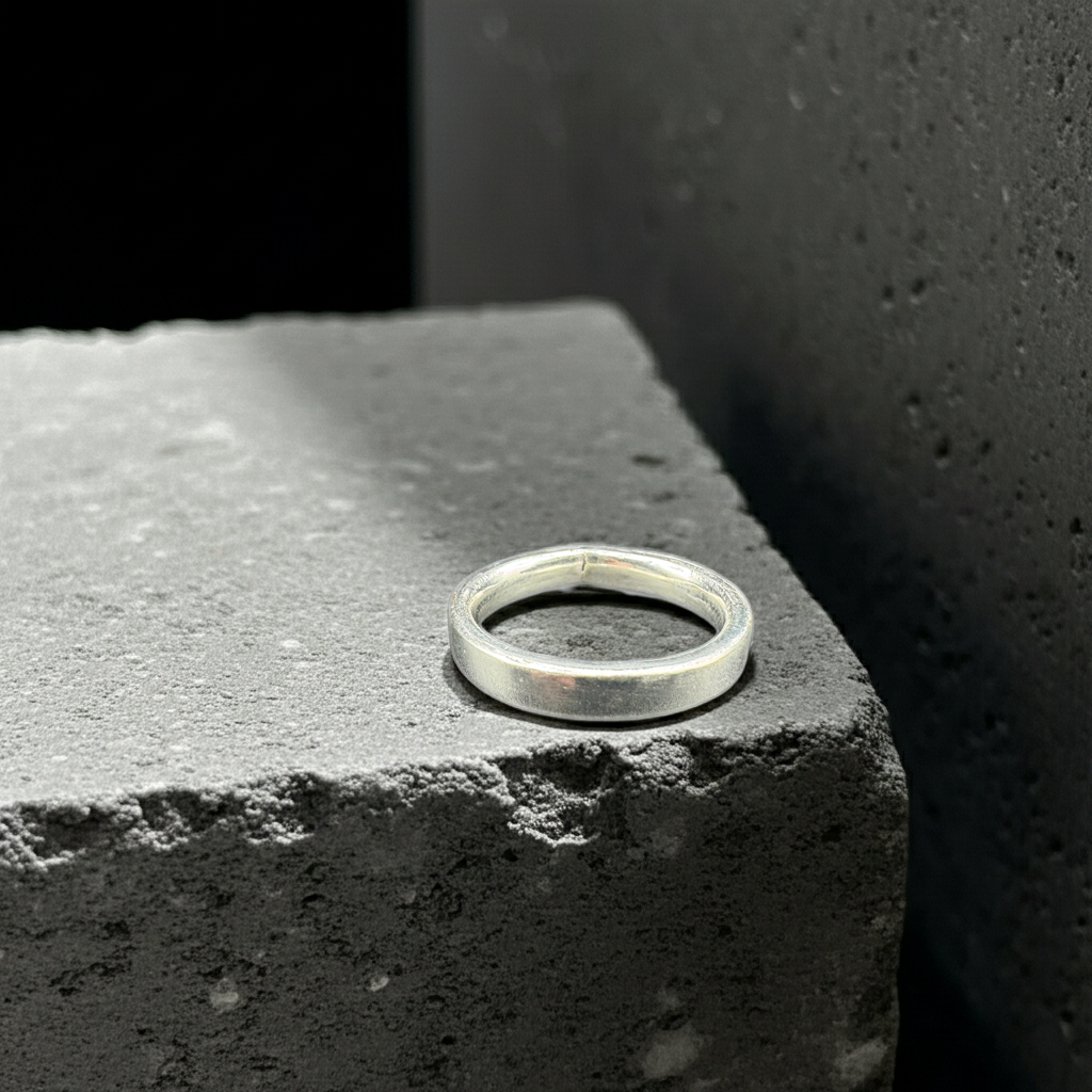 A minimalist silver band ring resting on a textured dark gray stone block against a dark background.