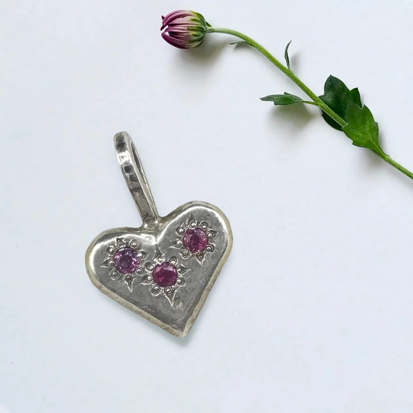 Handcrafted silver heart pendant with three purple gemstones beside a flower stem