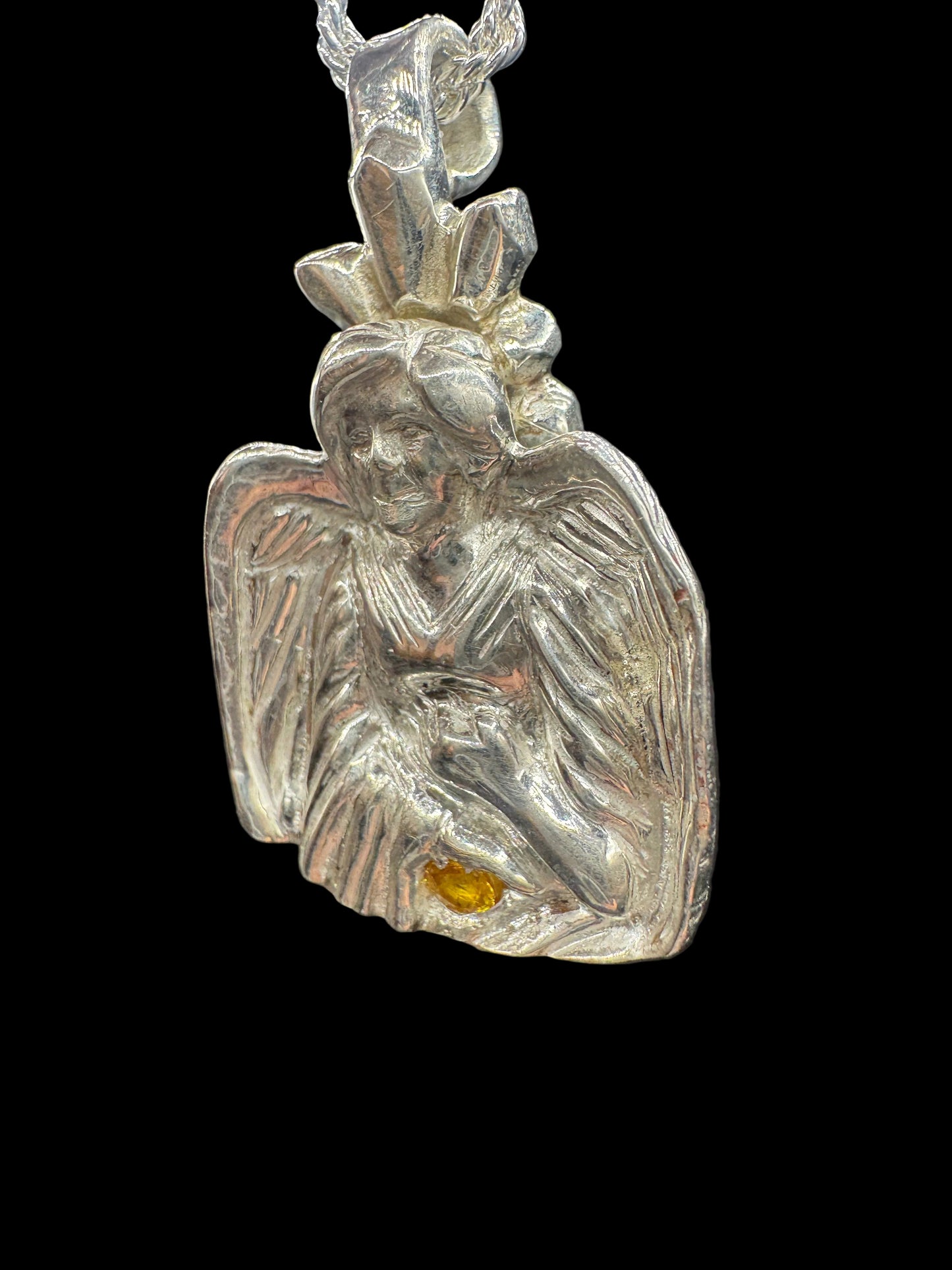 A silver-toned metal pendant or charm with a textured, organic leaf-like or wing-like shape, featuring a small amber-yellow inset detail, shown against a dark background.