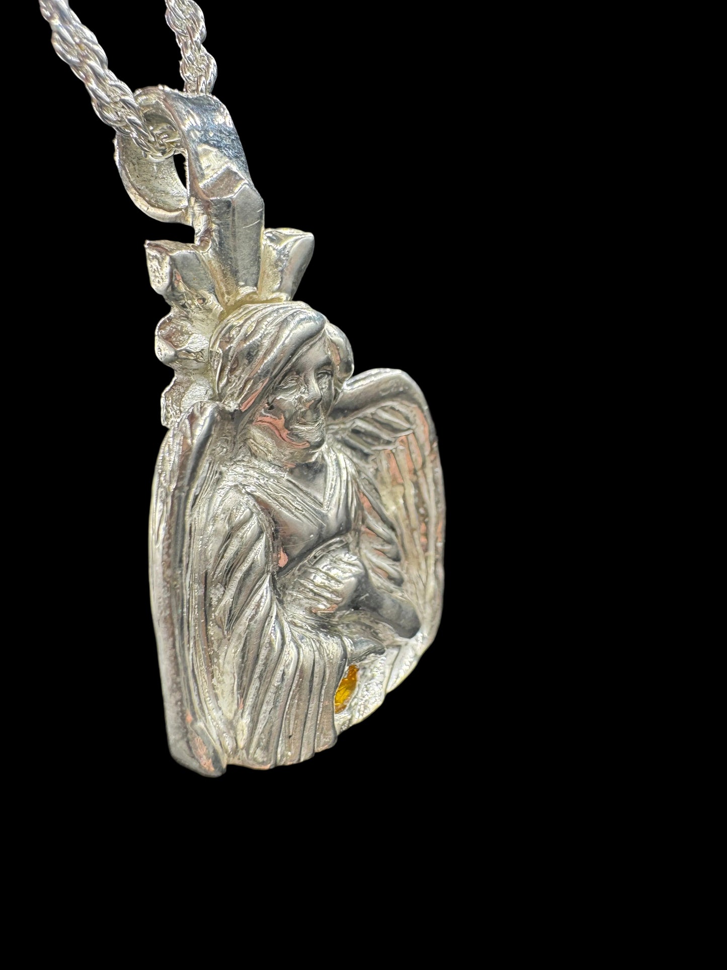 A close-up of a silver-toned pendant featuring a detailed angel or cherub design with wings and a bow, attached to a chain against a dark background.