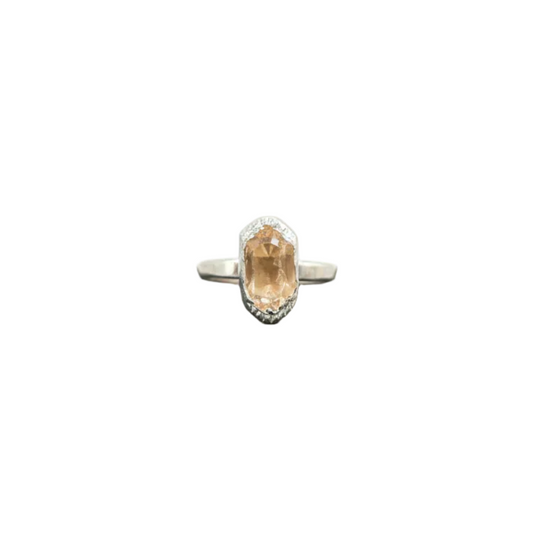 A silver ring with an oval peach-colored gemstone and small accent stones, displayed on a black slate stand against a white background.