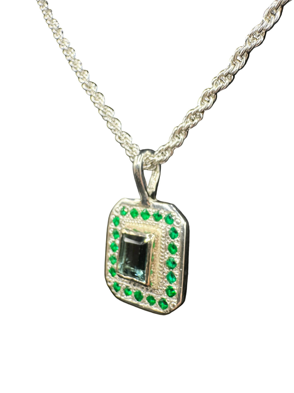 A silver-toned rectangular pendant necklace with a green rectangular center stone and a border of small green stones, displayed on a black background.