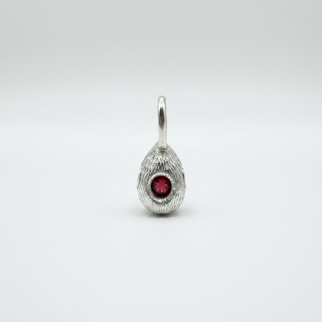 A silver teardrop pendant with a textured, feather-like outer surface and a central round red gemstone, shown on a dark gray background.