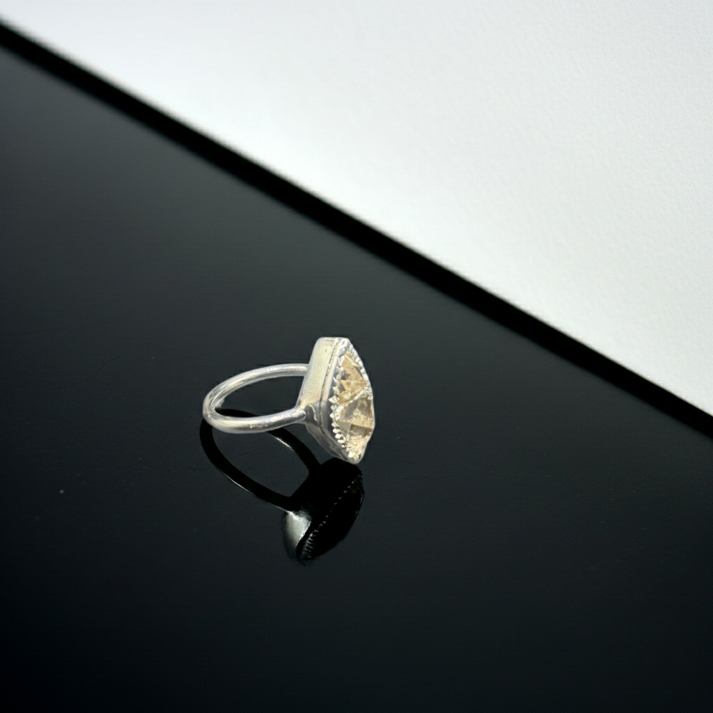 A silver ring with a triangular bezel-set clear gemstone, resting on a glossy black reflective surface with a white background in the upper right.