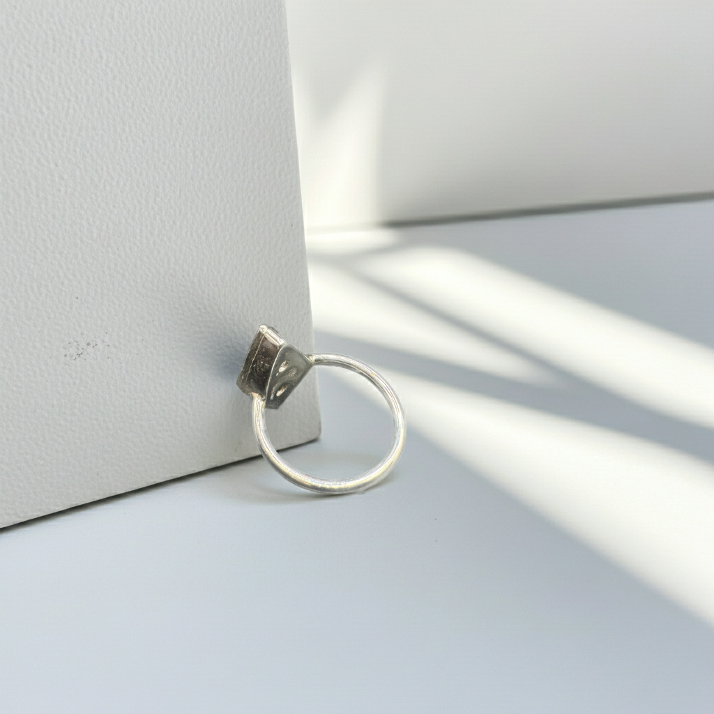 A minimalist silver ring with a textured, oxidized dark top and a thin circular band, resting on a light gray surface next to a white textured box under soft window shadows.
