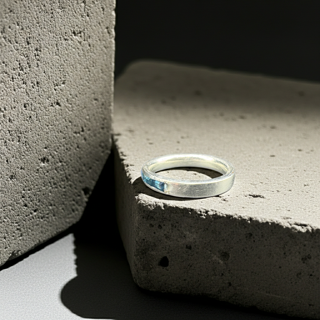 A polished silver band ring resting on a textured concrete block in dramatic, high-contrast studio lighting.