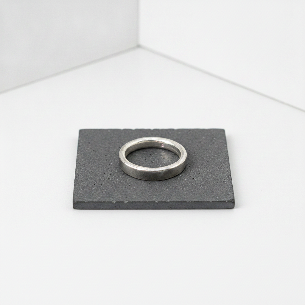 A minimalist silver band ring displayed on a square dark gray slate coaster against a clean white background.