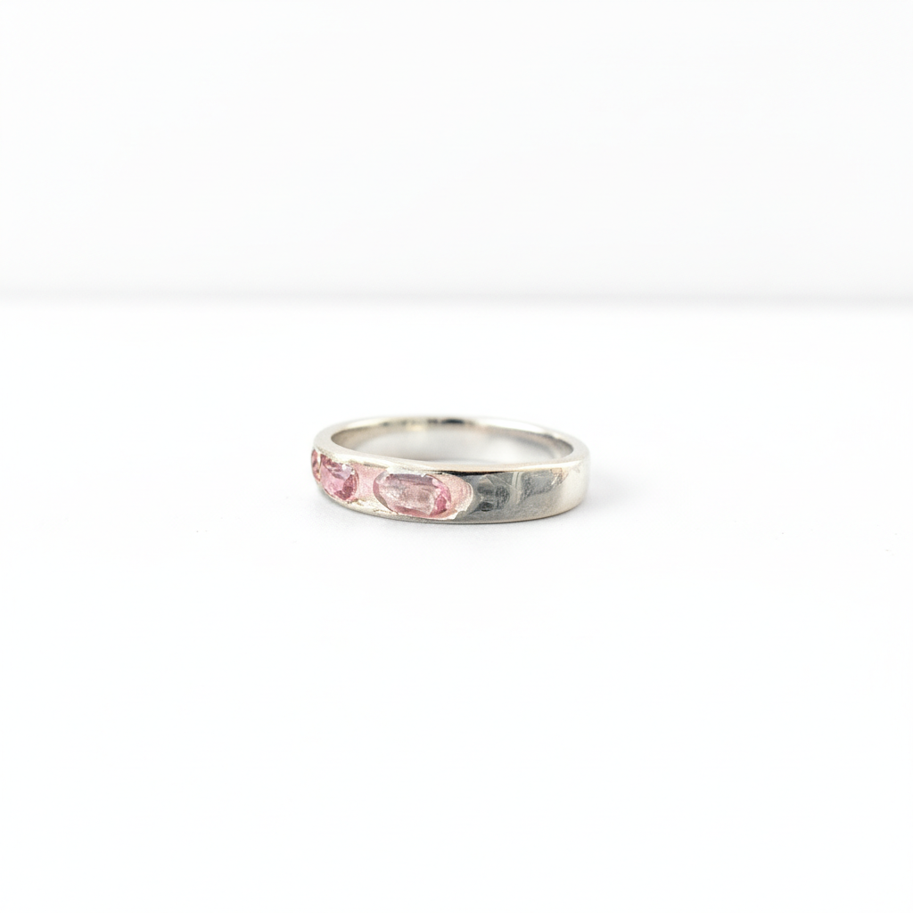 A silver band ring featuring three small, oval-cut pink gemstones set along the front, photographed on a seamless white background.