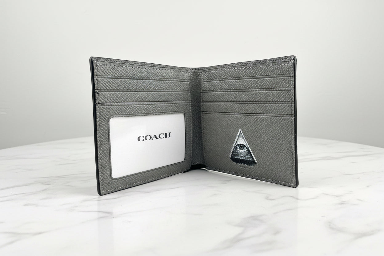 A dark gray textured leather bifold wallet shown open, featuring an ID slot with a white insert printed with the COACH logo and a small triangular emblem on the inside right panel, set on a white marble surface against a light gray background.