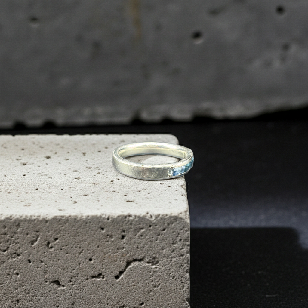 A silver band ring with a small light blue stone, resting on the corner of a textured light gray stone block against a dark background.